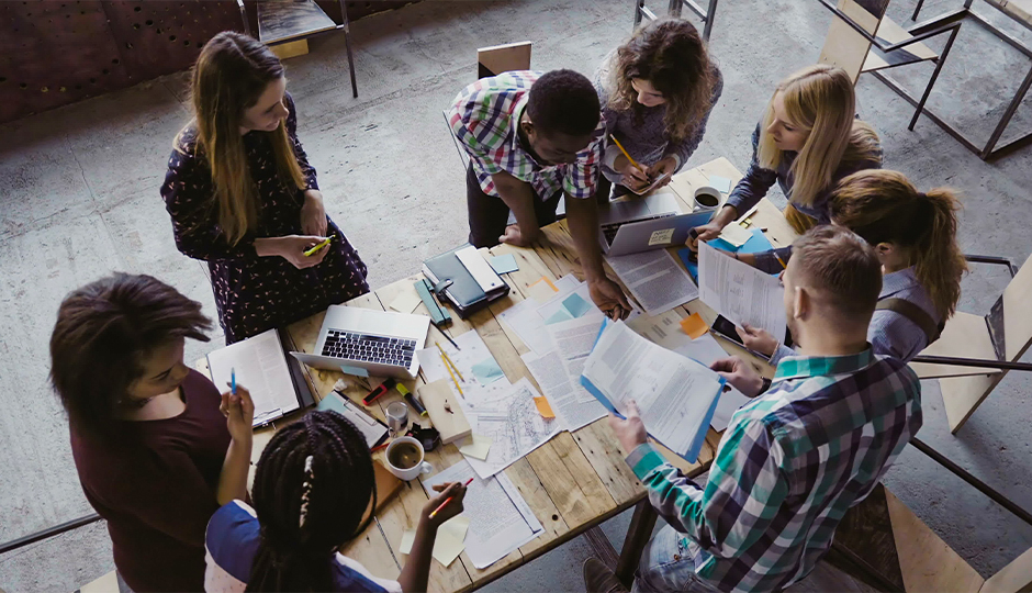 Eine Gruppe junger Menschen steht um einen Tisch mit Arbeitsmaterialien, Laptops, Blöcken, Stiften.