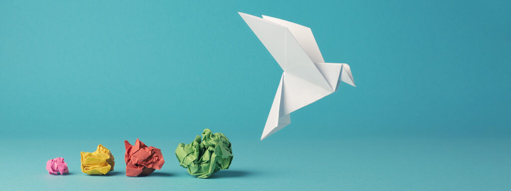 Fünf von links nach rechts größer werdende, farbige Papierknäuel. Rechts daneben ein weißer Origami-Vogel in fliegender Position.