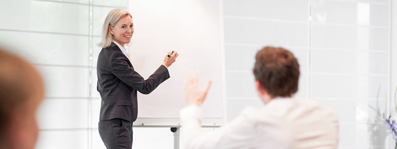 Eine Frau steht mit einem Filzstift neben einem Flipchart und blickt lächelnd nach vorn. Vor ihr sitzt ein Mann unscharf dargestellt und gestikuliert.