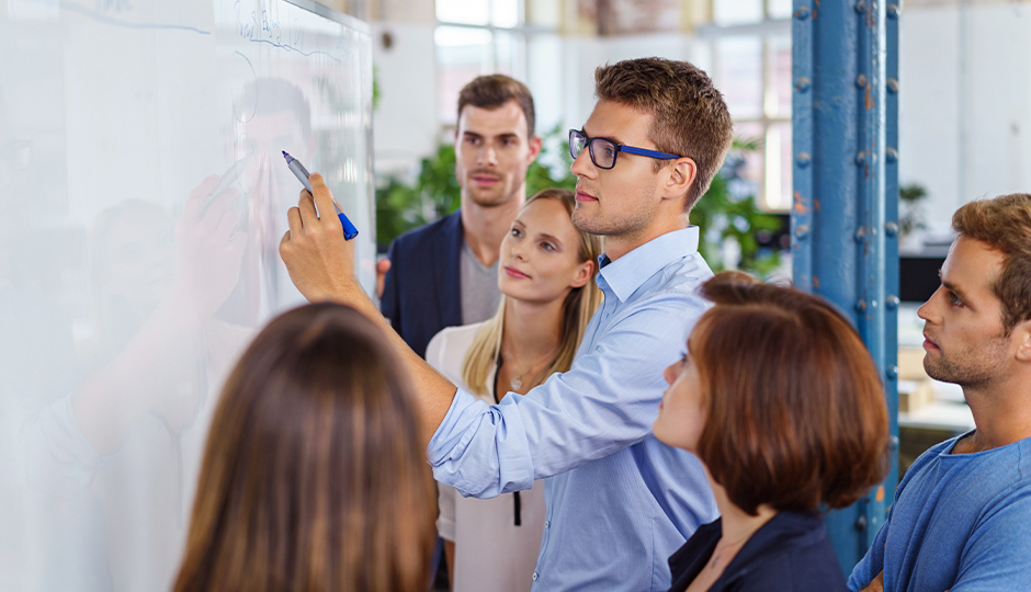 Ein junges Büroteam steht vor einem Whiteboard. Ein Mann setzt an, darauf zu schreiben. Fünf andere beobachten ihn dabei.