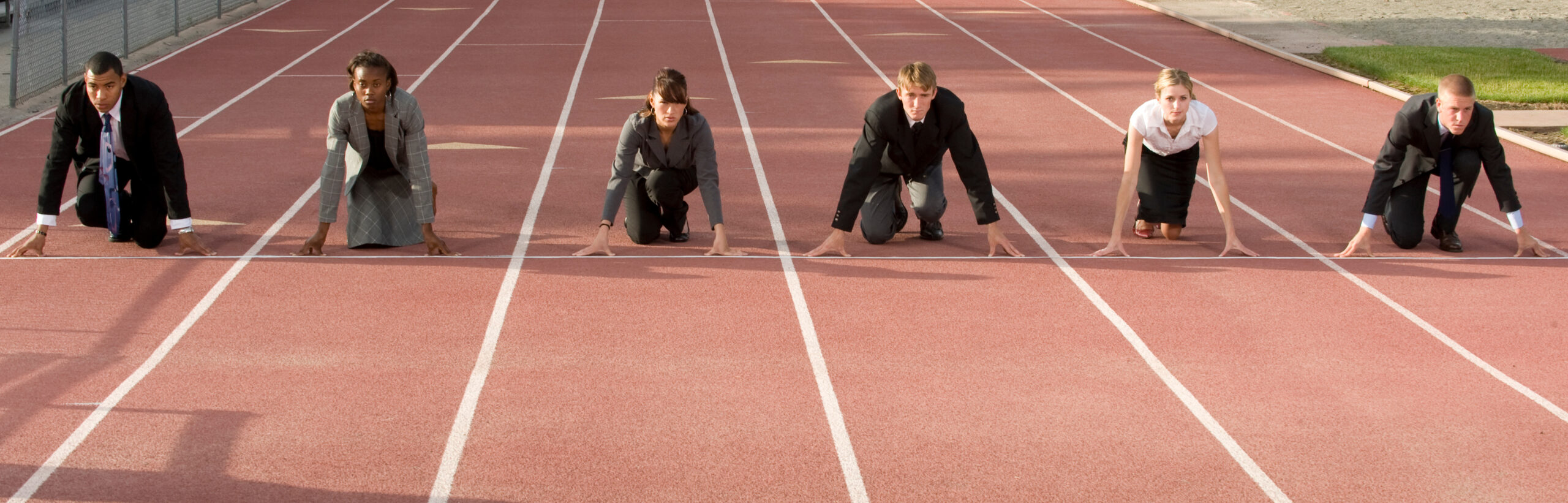 Sechs Menschen hocken im Business-Look an der Startlinie einer Rennbahn.