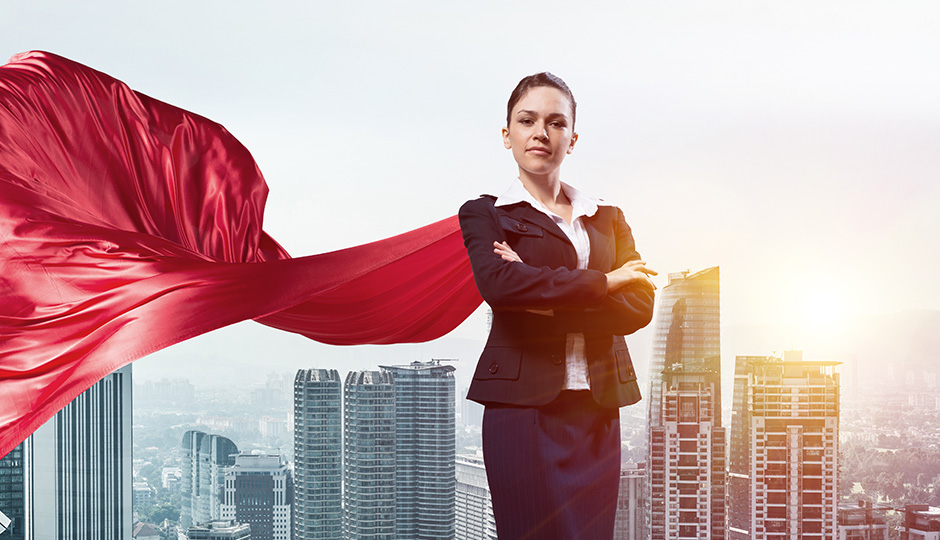 Eine Frau im Businesslook steht mit breiter Brust und verschränkten Armen hoch oben vor einer Großsstadt. Sie trägt eine rotes Superhelden-Cape am Rücken.