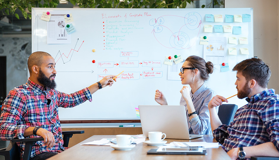 3 Menschen sitzen mit Laptops und Kaffeetassen an einem Tisch und unterhalten sich. Im Hintergrund ein beschriebenes und mit Klebezetteln beklebtes Whiteboard.